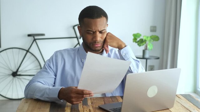 Pensive and worried young african american businessman reading a document with bad news, sitting at his desk at home office with a laptop, looking frustrated about his work problems
