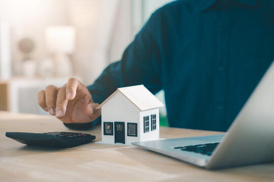 Investment planning for real estate involves man using calculator next to house model and laptop for home mortgage loan, calculating property tax and managing insurance and investment assets