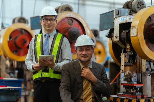Two workers stand in a factory with machines while one holds a tablet and the other adjusts his tie during a workday
