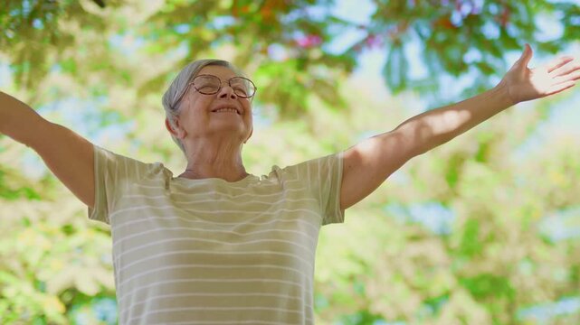Elderly woman with glasses smiling and breathing fresh air in a park, feeling free and enjoying a beautiful sunny day with her arms outstretched in a gesture of pure happiness