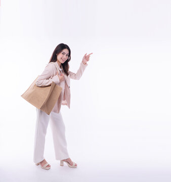 Smiling young Asian woman in beige suit carrying eco friendly jute shopping bag, pointing finger at empty copy space on white background. Sustainable retail shopper for green marketing.