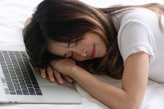 Close up of a tired but happy young Asian woman sleeping peacefully on her hands next to a laptop on a white bed. Relaxed female freelancer taking a nap after working or studying at home.