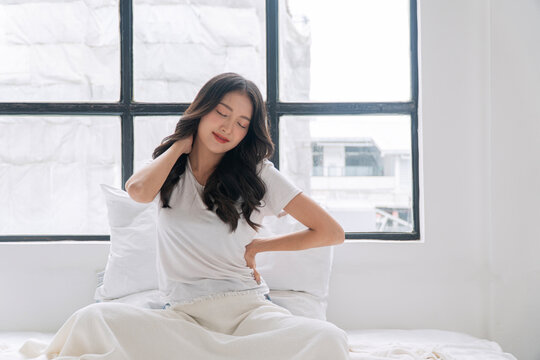 Young Asian woman in white t-shirt sitting on bed by the window, stretching neck and massaging back with closed eyes. Relaxed female waking up or relieving muscle tension in cozy bedroom.
