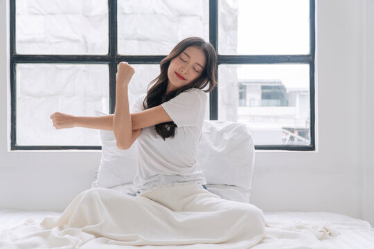 Smiling young Asian woman in white t-shirt stretching arms while sitting on a cozy bed by the window in morning. Relaxed female waking up feeling fresh and rested in bright bedroom.