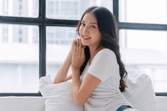 Smiling young Asian woman in white shirt sitting on bed by large window. Happy beautiful female resting in bright cozy bedroom, looking at camera. Relaxed morning lifestyle and wellness concept.