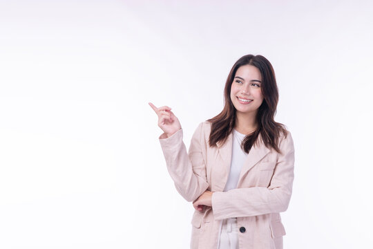 Smiling young Asian businesswoman in beige suit pointing finger at empty copy space on left over white background. Cheerful female professional recommending product for corporate advertising.