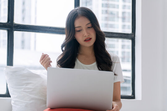 Stressed young Asian woman working on laptop at home by the window. Frustrated female freelancer having internet problem, reading bad news or facing difficult task in bright bedroom.