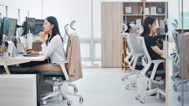 A wide view of a young professional woman in a beige blazer at a workstation in a bright modern open-plan office with city skyline visible.