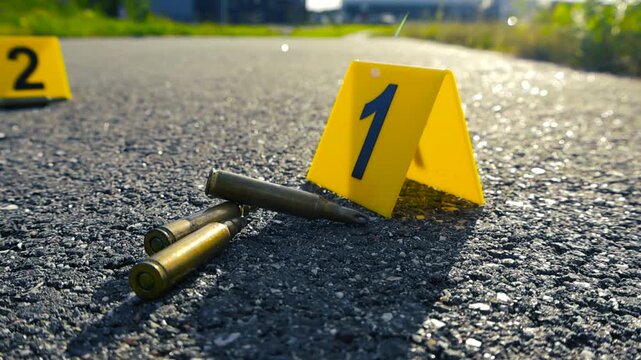Yellow forensic evidence markers numbering the bullet casings found on the asphalt road at a crime scene, suggesting a recent shooting and ongoing police investigation into a violent crime