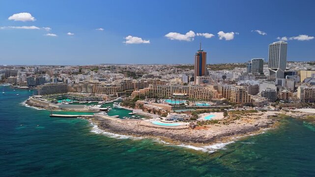 St Julian's Bay in Malta with Portomaso and Mercury towers above the marina, yachts and turquoise Mediterranean coastline under a blue sky in an aerial cityscape view.