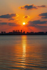 Fototapeta premium Bright sunset glows over city skyline near water. Golden light reflects on lake surface during evening. Urban buildings create black silhouette against orange sky horizon. Nature landscape view