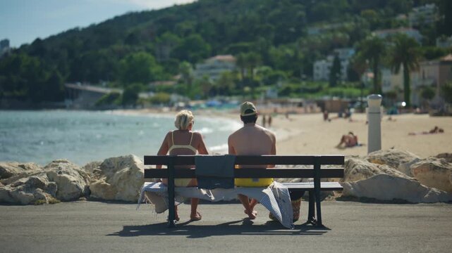 Couple sitting on a bench facing the sea and beach, with a sunny coastline and green hillside in the background