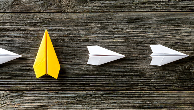 The yellow paper airplane flies ahead of the white ones on the wood.