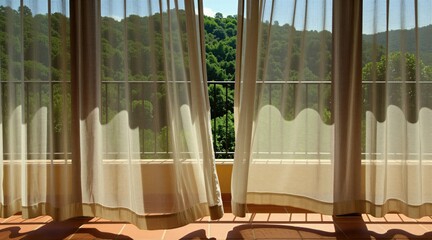 Obraz premium Bright golden sunlight streams through sheer beige curtains onto rustic tiled patio. Transparent fabric drapes flutter softly near open glass door revealing green forest view outside. Warm morning