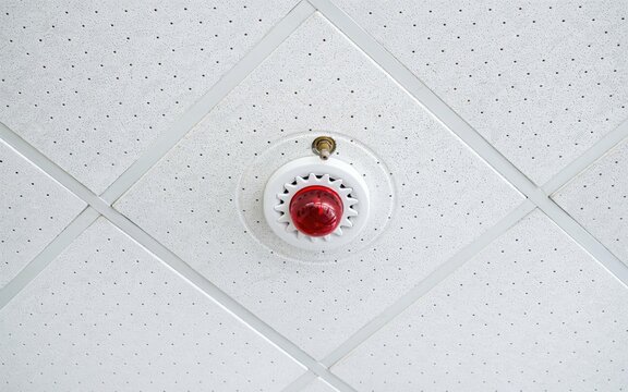 Bright red fire alarm strobe light mounted on a white acoustic tile office ceiling panel