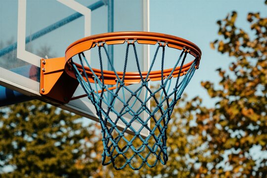 Close-up of outdoor basketball hoop net with orange rim, blue ropes. Transparent backboard visible, with blurred autumn trees in background under clear sky. Image perfect for sports recreation themes.