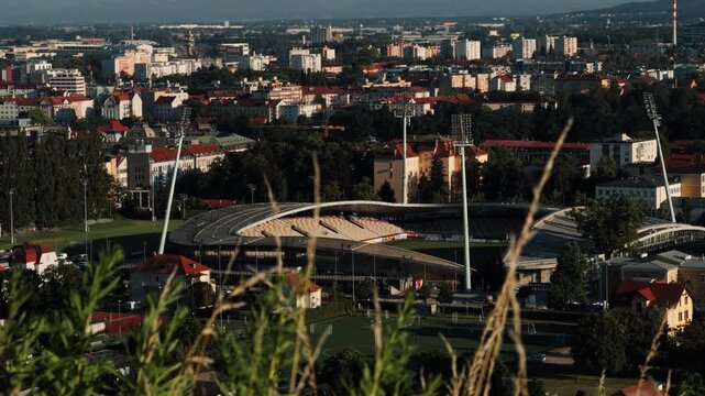 Maribor from a hillside, showing the football stadium surrounded by city buildings, red rooftops, trees, sports fields, and distant hills.