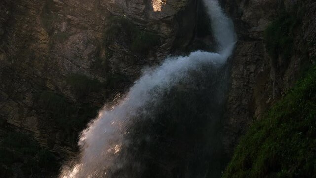 A powerful waterfall rushes through a rocky gorge, sending spray across dark stone walls with patches of grass and sunlight catching the fast current.
