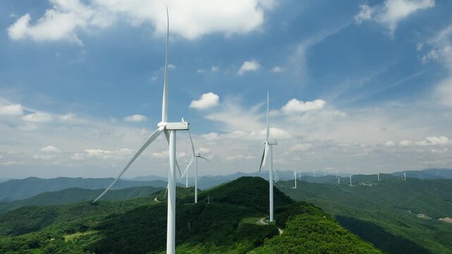 Aerial zoom out timelapse of a wind turbine farm on a forested mountain ridge with fast-moving clouds against a blue sky. Wind turbines generate clean, renewable energy. Yeongnam Alps, South Korea.