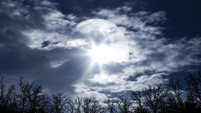 Dramatic night sky with a large full moon partially obscured by clouds silhouetted trees in foreground stars visible with video captures dramatic moon peeking