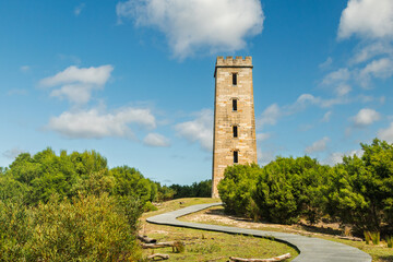 Fototapeta premium A curved boardwalk pathway leads up to Boyd's Tower, a rejected lighthouse and now a landmark ruin set in lush coastal bush, against a blue sky with clouds in Beowa National Park near Eden, Australia.