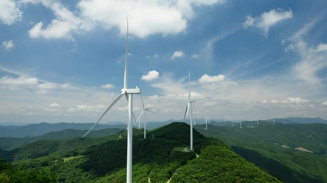 Aerial timelapse of a wind turbine farm on a forested mountain ridge with fast-moving clouds against a blue sky. Wind turbines generate clean, renewable energy. Yeongnam Alps, South Korea.