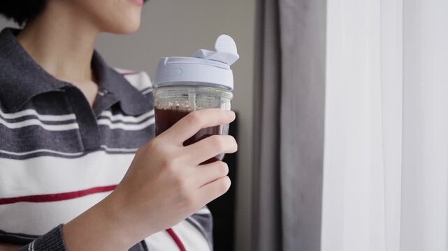 Young woman sips cold brew coffee from a tumbler glass, enjoying a quiet morning moment at home.