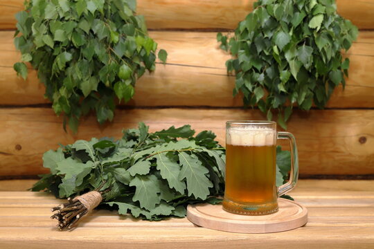 A mug of light beer on a round wooden tray next to a fresh oak broom stands on a plank bench against the background of a log wall with a couple of fresh birch brooms hang on it.