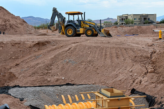 Stormwater drainage infrastructure under installation at a commercial development site with a backhoe loader in the background, representing utility construction, excavation, and site preparation