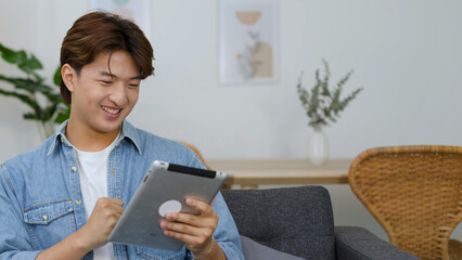 Excited Young Asian Man Celebrating Success While Using Digital Tablet on Sofa © blueringmedia
