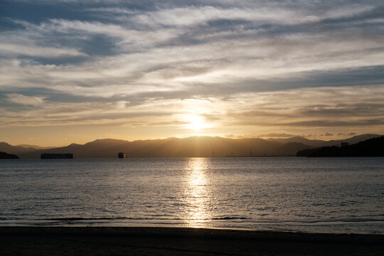 Golden sun dips over calm waters. Sky glows with soft amber hues. Clouds stretch wide across horizon. 