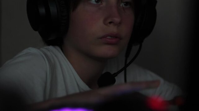 A close-up shot of a young gamer's face in headphones with a microphone during a night gaming session. The teenager intently follows the online match