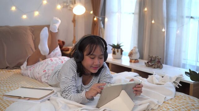 Relaxing Evening, Young girl wearing headphones lies comfortably on bed using a tablet in a warm, cozy bedroom decorated with soft fairy lights