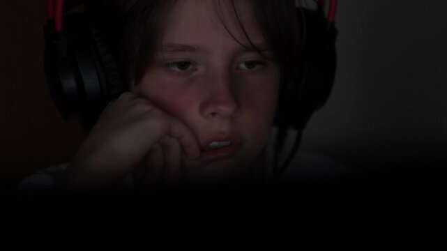 A tired teenager in large gaming headphones props up his face with his hand in front of a computer screen. The young gamer looks sad or bored during a late-night session in a dark room.