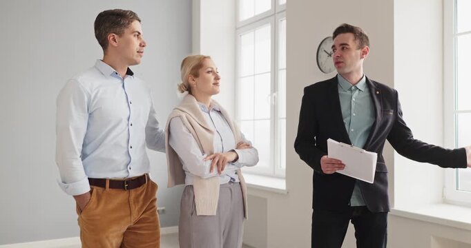 Realtor guides couple clients on apartment tour. Agent presents window features and layout in a bright empty space, discussing purchase, sale or rent. Modern home search and choices made.