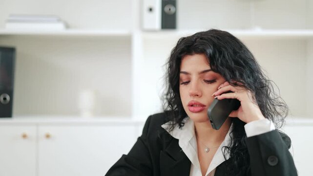 Professional woman with dark, curly hair engages in serious phone conversation. Her expression conveys concentration and concern while discussing important matters in an office. Communication is key.
