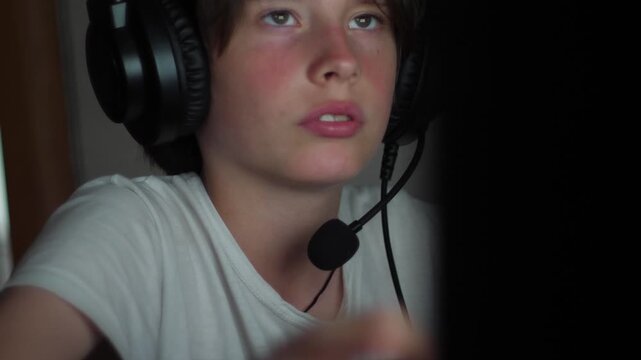  The young gamer's face is periodically illuminated by bright flashes from an unseen monitor in a dark room