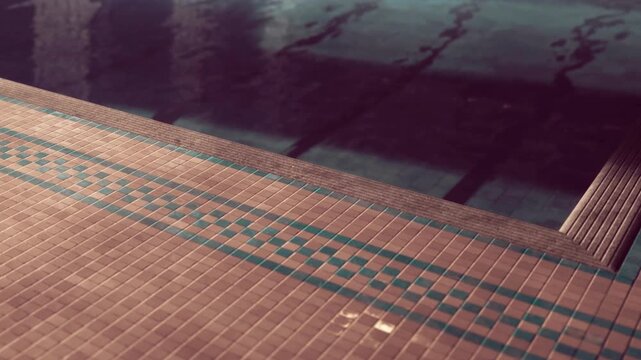 Water reflects light on the quiet pool surface. The edge of the pool has tile patterns and borders. The scene shows a moment in a swimming area, likely in a recreational facility.