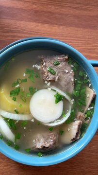 A close-up overhead shot shows a blue bowl of traditional Bukhler soup with beef ribs, onion, and herbs on a wooden table