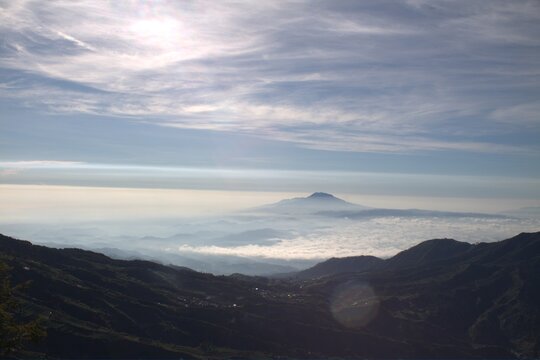Majestic mountain peak rising above clouds under bright hazy sun. Panoramic volcanic landscape with silhouetted ridges and ethereal morning light.