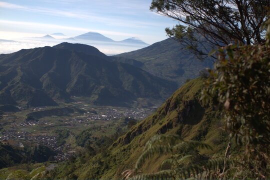 Panoramic mountain valley view with village, distant volcanic peaks above clouds, bright morning light, lush green slopes.