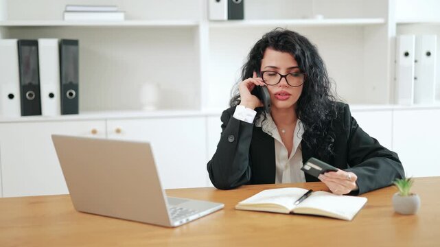 Angry businesswoman holding credit card calling bank support on phone, complaining about missing money or transaction issue. Stressful office situation, financial problem concept in modern workspace