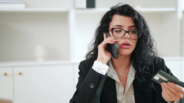 Frustrated businesswoman holding credit card and smartphone, calling technical support to complain about missing money or unauthorized transaction. Stressed financial situation in modern office