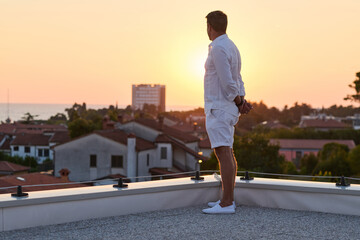 Obraz premium Man Standing on Rooftop Terrace Watching Sunset Over Seaside Town From Home Balcony