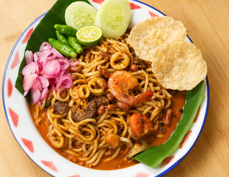 Traditional Indonesian Mie Aceh with seafood, beef, spicy curry broth, fresh cucumber, lime, pickled shallots, chilies, and melinjo crackers served on a classic plate.