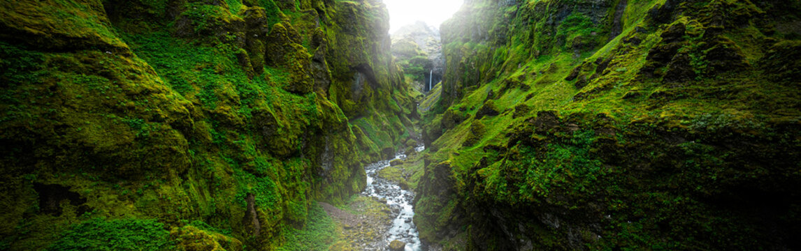 Panoramic view of the breathtaking M&uacute;laglj&uacute;fur Canyon in southeast Iceland, featuring dramatic moss-covered cliffs, a cascading waterfall, and a winding river beneath soft mountain light.