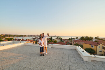Obraz premium Family Taking Selfie On Rooftop Terrace Overlooking Coastal Town And Sea At Sunset