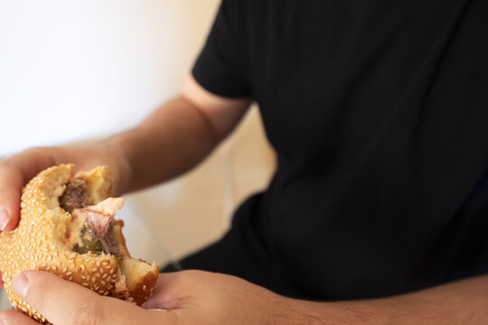 Man holding a sesame seed burger with a bite taken out, showcasing delicious layers of flavor in a cozy setting.