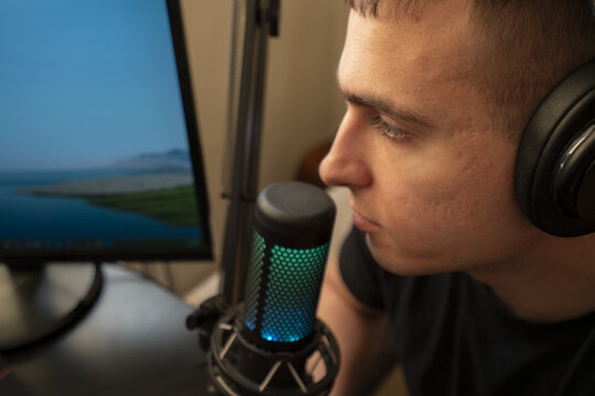 Young man speaking into a microphone while working at a desk with a computer screen in the background, ideal for content creation or podcasting.