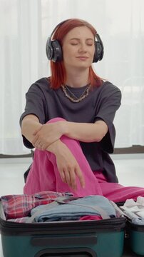 Vertical Screen: A woman sits in a room with her suitcase open. She listens to music through headphones. Clothes and money in euros are nearby. She looks relaxed and lost in thought.
